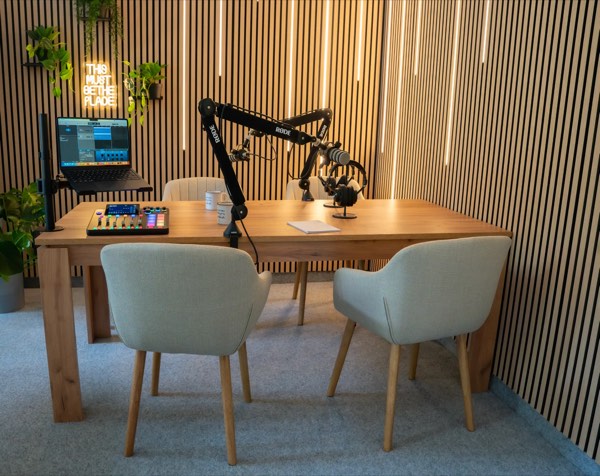 Studio interior: wooden table, four armchairs, microphones on RØDE boom arms and a 'This must be the place' neon